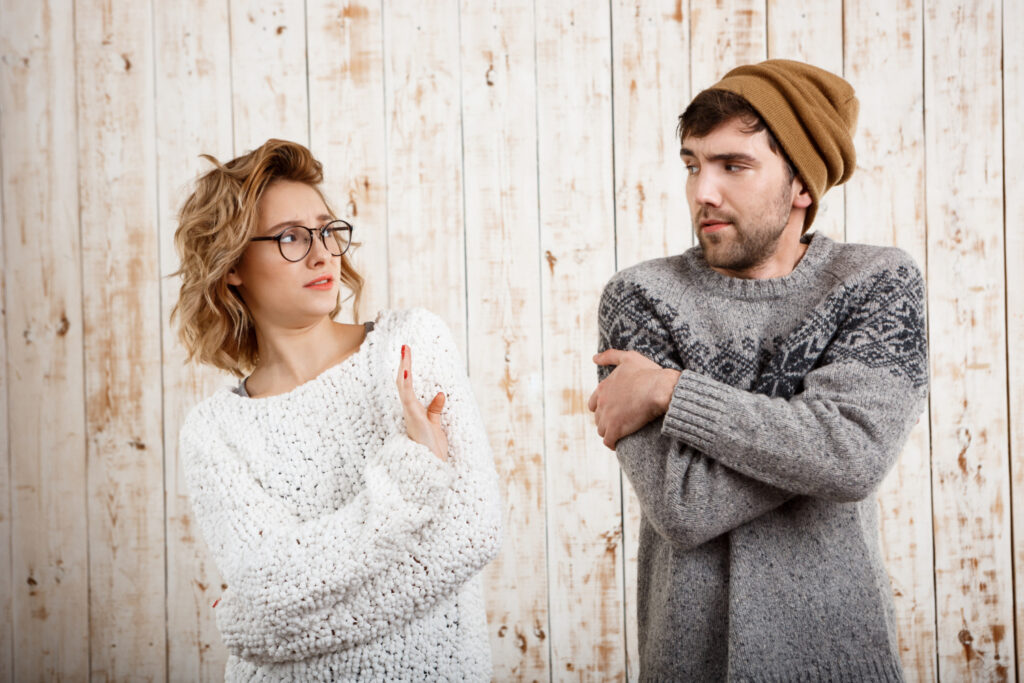 Ein Mann mit Bart und Mütze in einem grauen Pullover verschränkt die Arme und blickt nach links zu einer Frau mit Brille und gewelltem Haar, die einen weißen Pullover trägt und eine Hand hebt.