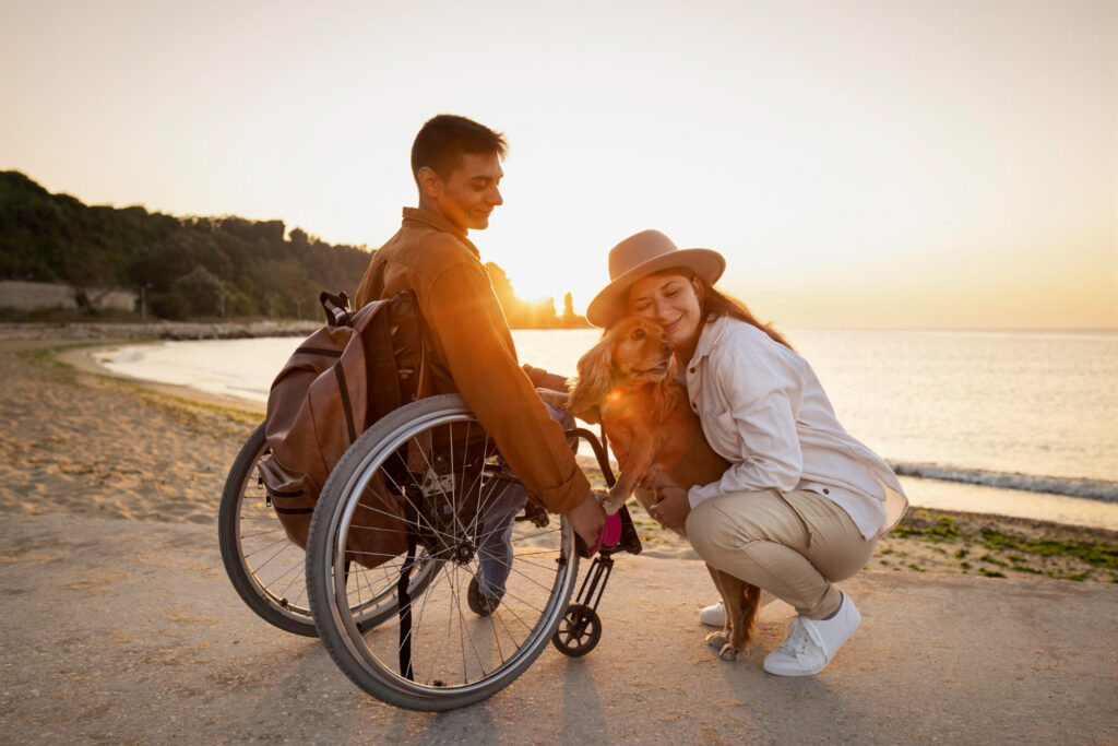 Eine Frau hockt neben einem Mann im Rollstuhl am Strand, die Sonne geht über dem Meer unter und wirft einen goldenen Schein auf die Szene.