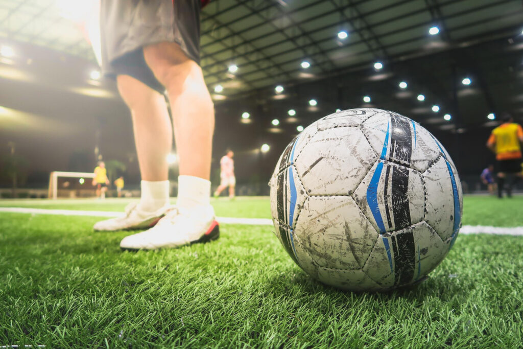 Ein abgenutzter schwarz-weiß-blauer Fußball liegt auf einem grünen Rasenfeld, im Hintergrund sind die Beine eines Spielers in weißen Schuhen und verschwommene Scheinwerfer zu sehen.